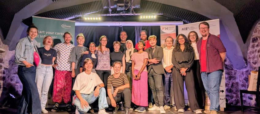 Duża, uśmiechnięta grupa ok. 20 osób pozuje na scenie w kamiennej sali z łukowym sufitem; w tle roll-upy partnerów, m.in. Infineon i ACE² European University, sceniczne światła i sprzęt.